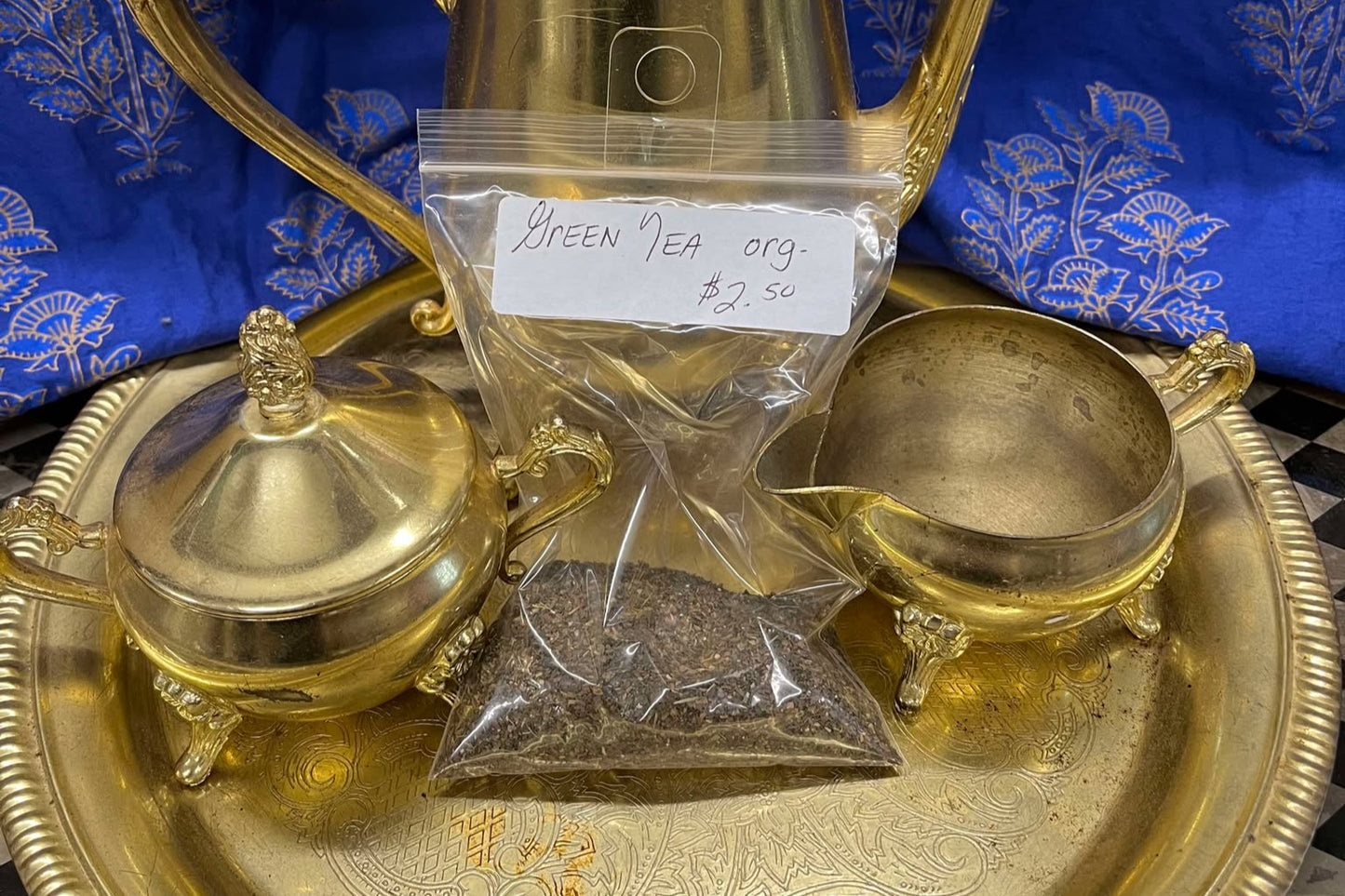 Green, brown and tan tea leaves in a clear plastic zip bag sit on a golden tea tray in surrounded by a tea set. The bag is labeled "Green tea, organic, $2.50 per oz"