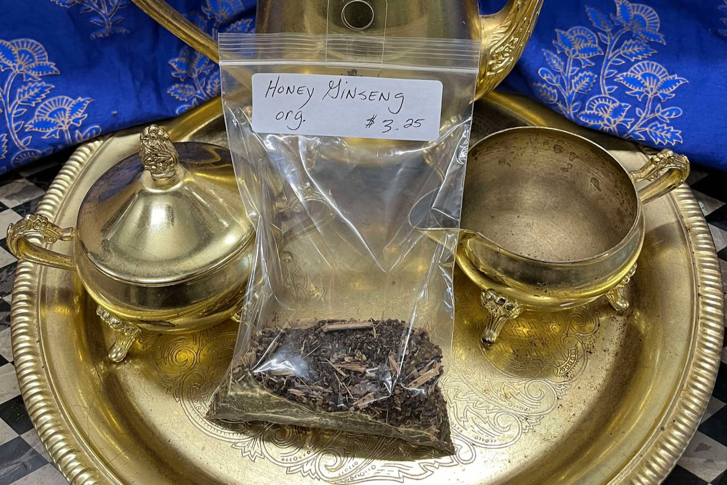 Dried tea leaves infused with small pieces of thin wood in a clear plastic zip bag sit on a golden tea tray surrounded by a tea set. The bag is labeled "Honey Ginger Tea, Organic, $3.25 per Oz