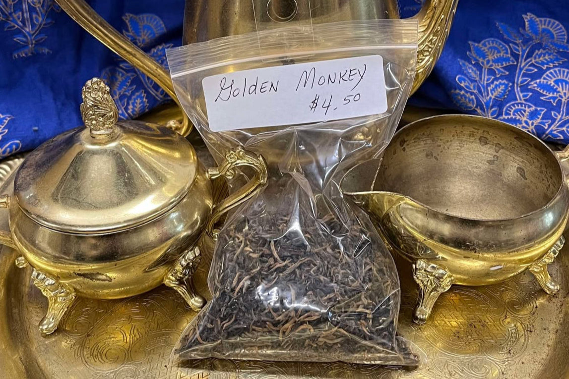 Brown and tan dried tea leaves in a clear plastic zip bag sit on a golden tea tray surrounded by a tea set. The bag is labeled "Golden Monkey Tea, $4.50 per Oz