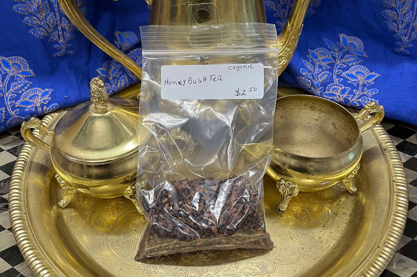 Redish/brown small pieces of thin wood in a clear plastic zip bag sit on a golden tea tray surrounded by a tea set. The bag is labeled "Honeybush Tea, Organic, $2.50 per Oz