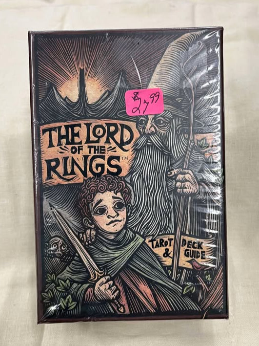 drawing of a wizard (gandalf), a hobbit (Frodo) holding a sword, with the sun setting behind a mysterious tower in the background. The deck reads "The Lord of the Rings: Tarot Deck & Guide."