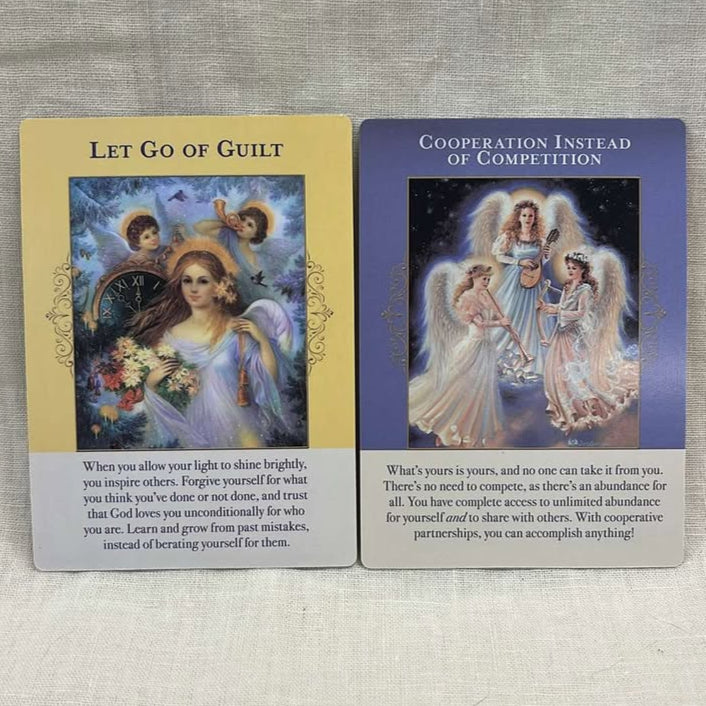 Two angel-themed cards on a textured surface. One card says "Let Go of Guilt" with one large angel and two small angels on a yellow background with text beneath. The other card says "cooperation instead of competition" with three angels together in the night sky, each playing a different instrument (horn, harp, guitar) on a blue background with text below.