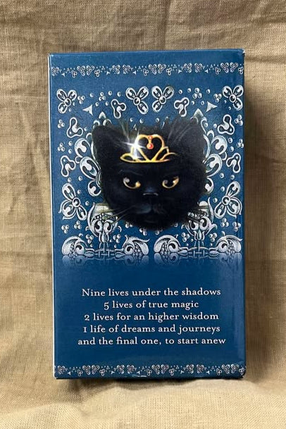 Blue card deck back with a black cat wearing a golden crown on a blue background with a white design. Below are the words "Nine lives under the shadows, 5 lives of true magic, 2 lives for an higher wisdom, 1 life of dreams and journeys and the final one, to start anew."