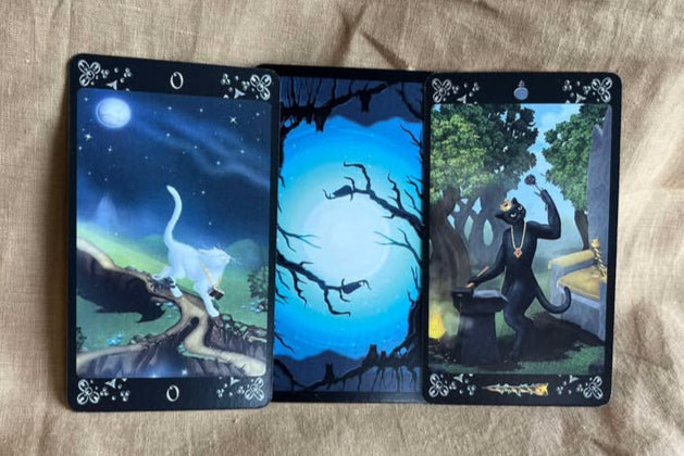 Three tarot cards with nature-themed illustrations on a textured fabric background. The first has a white cat walking along a moonlit path, the second has a card back featuring tree branches in front of a full moon, and the third has a black cat blacksmithing a metal rose at a forge.