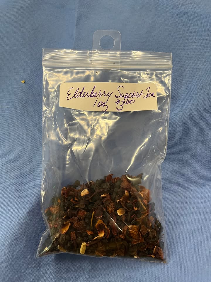 Blend of red dried rose hips, black dried elderberries, dark red dried flowers, and other dried herbs in a clear plastic bag, with a label saying "Elderberry Support Tea, 1 oz for $3.00"