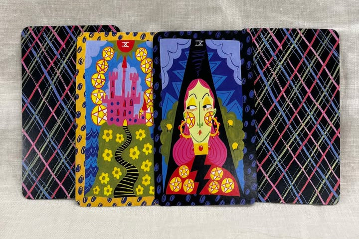 Colorful tarot cards with artistic designs on a textured surface. The card backs have diagonal stripes of white, blue, pink going both directions.