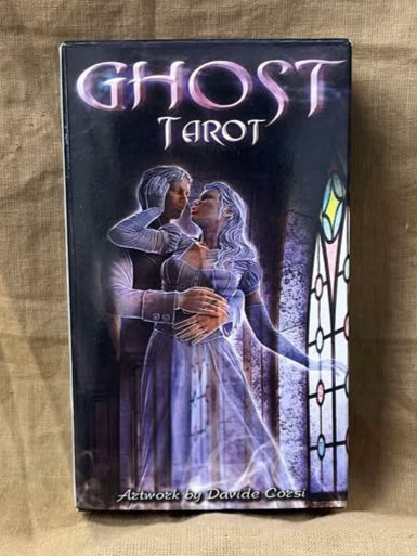Ghost Tarot card deck packaging on a textured surface. A man and woman, transluscent in spirit form, stand before a stained glass window. The man is slightly behind the woman and has his hands around her waist. The woman has turned to kiss the man, with her arm gently caressing the side of his face.