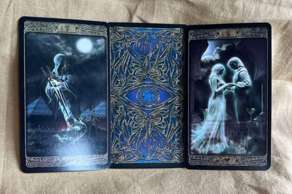 Three tarot cards with mystical designs on a textured fabric background. The first has a robed spirit clutching four swords to its chest, in front of a camp where two swords are propped up . The second shows the card backs, a blue background with silver etching with an eye in the middle. The third card shows two lover spirits set against a moonlit night, on a balcony. The male holds the hands of the female as they face one another.