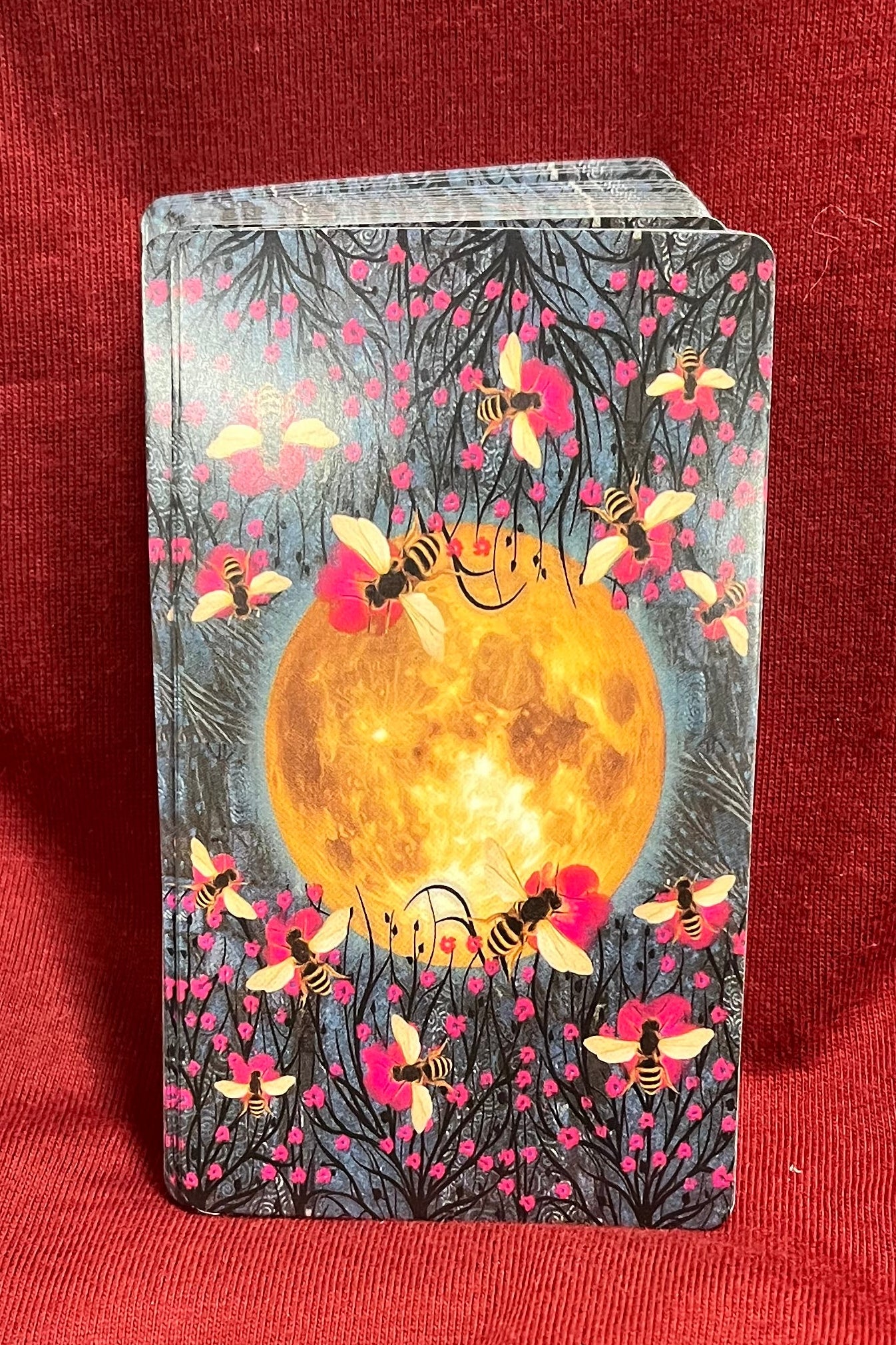 A deck of illustrated cards standing upright on a red fabric background. The card back features a large glowing yellow moon surrounded by pink flowers, bees, and dark tree branches.