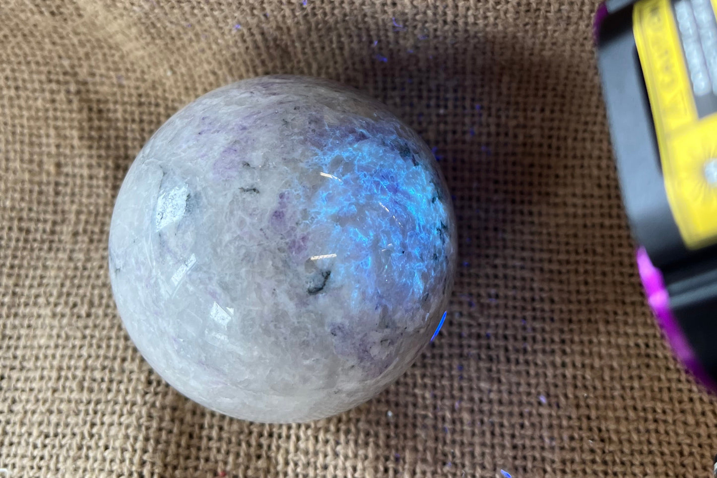 Polished white and light purple crystal sphere with darker purple marbling on a textured beige background. A black light is pointed at the stone, which makes parts of it glow an electric blue and make other parts of the purple darken.