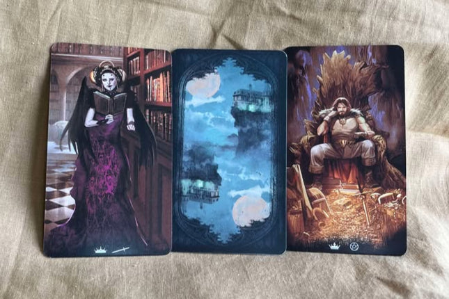 Three tarot cards on a textured surface. One has a woman in a purple and black dress, with black wings and an intricate hairstyle, reading a book in a library. The next is a card back featuring a moonlit scene of a cliff over water. The last features a man, sitting on a throne, surrounded by golden treasures.