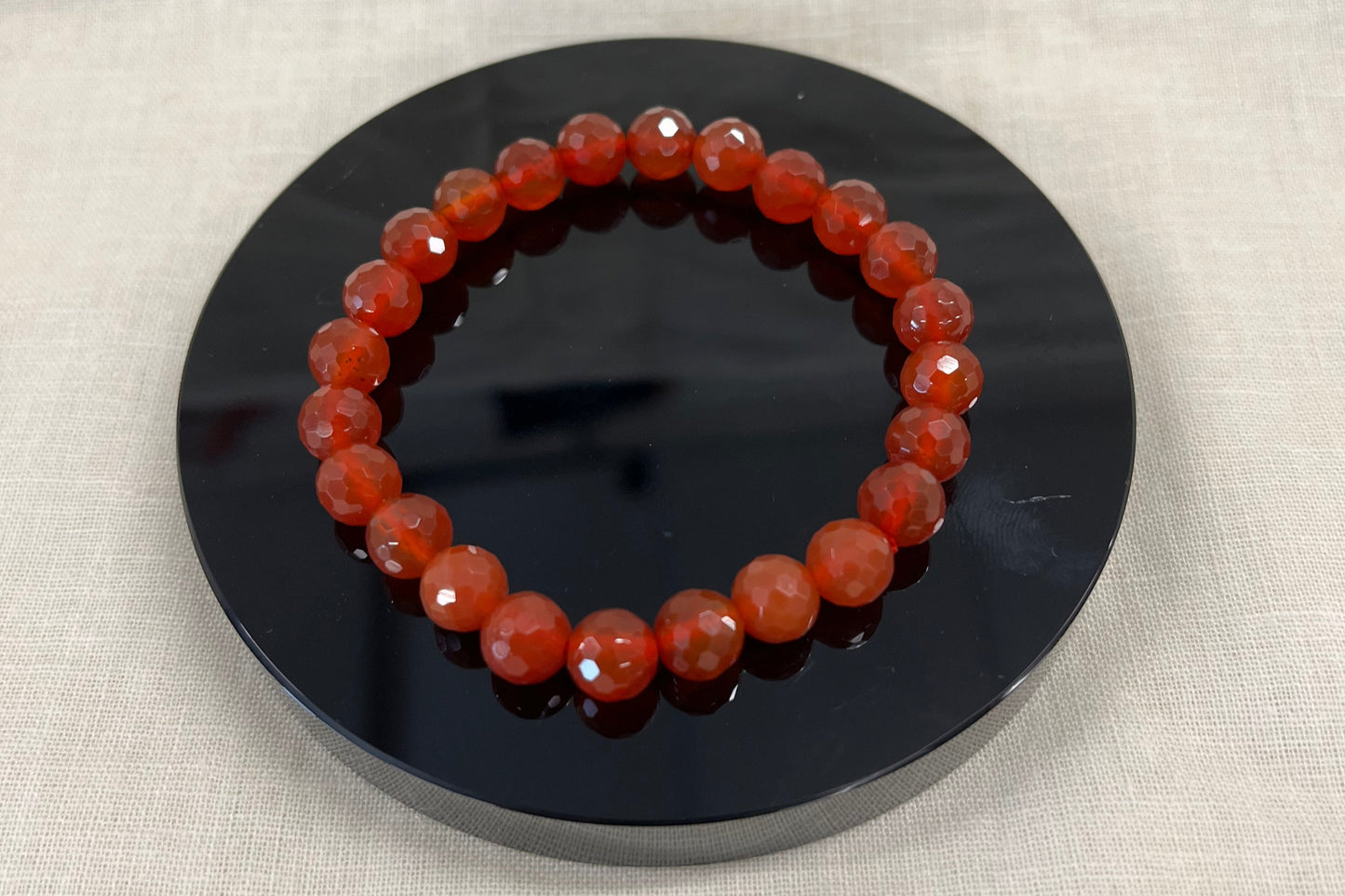 Redish orange faceted carnelian bead bracelet on a black circular stand against a beige fabric background