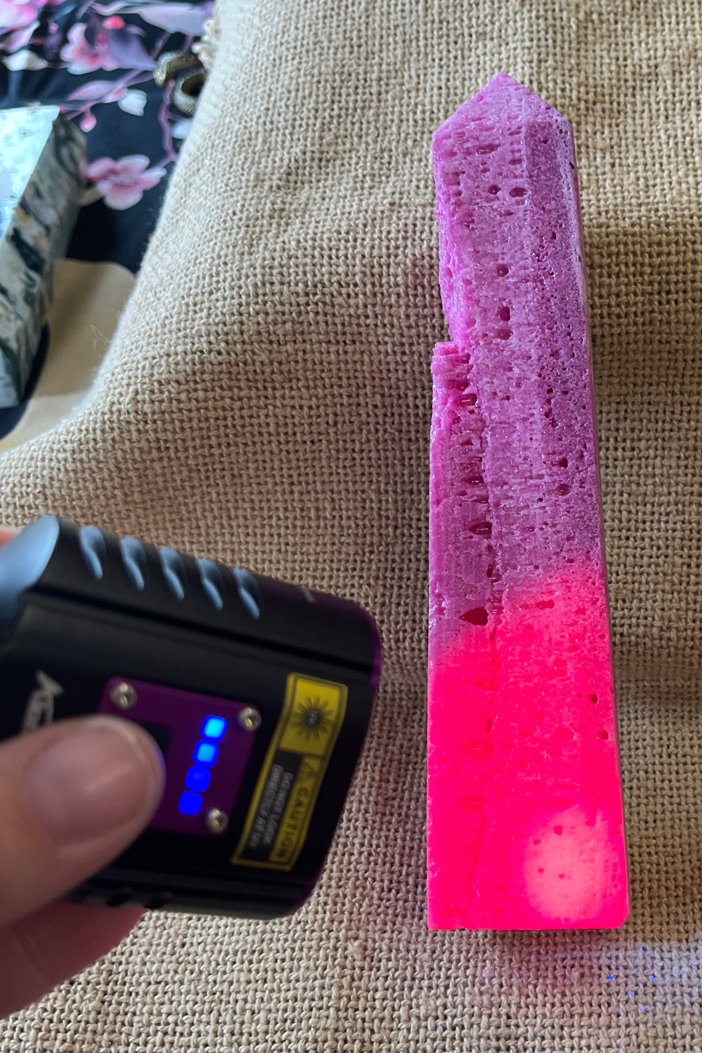 Purple-ish pink unpolished crystal tower on a textured surface with a black light held above it. The areas hit by the black light glow a much more vibrant pink color.
