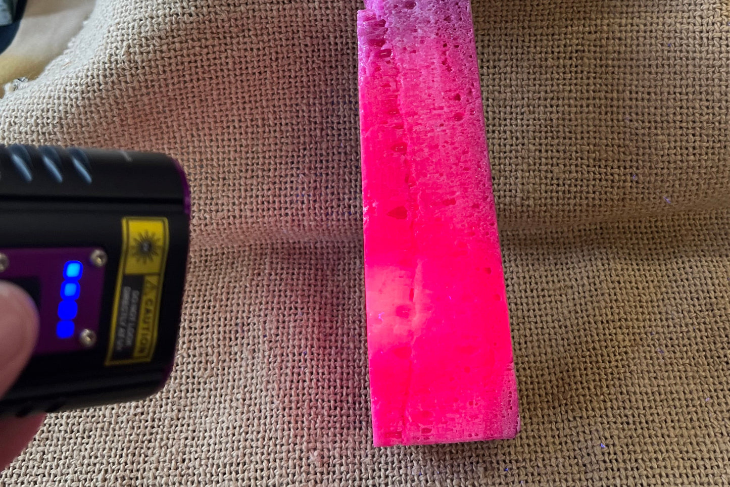 Purple-ish pink unpolished stone tower on a burlap background. A black light flashlight is pointed toward the crystal, and the areas where the light hits are a much more vibrant pink color