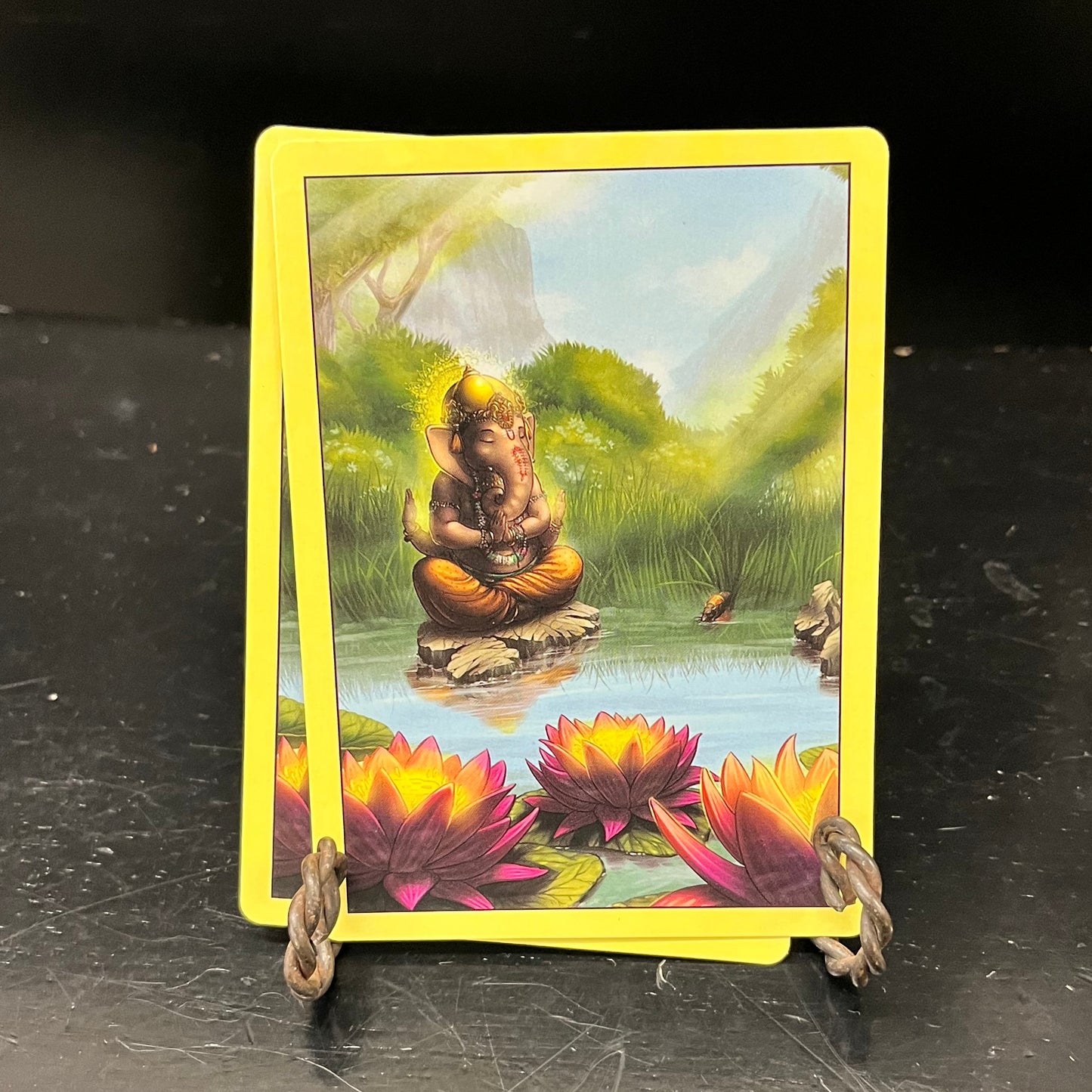 Card backs shows an elephant, in clothing and jewelry, meditating on a rock in the middle of a pond with flowering woods behind it. Glowing lotus flowers float on the surface of the pond.