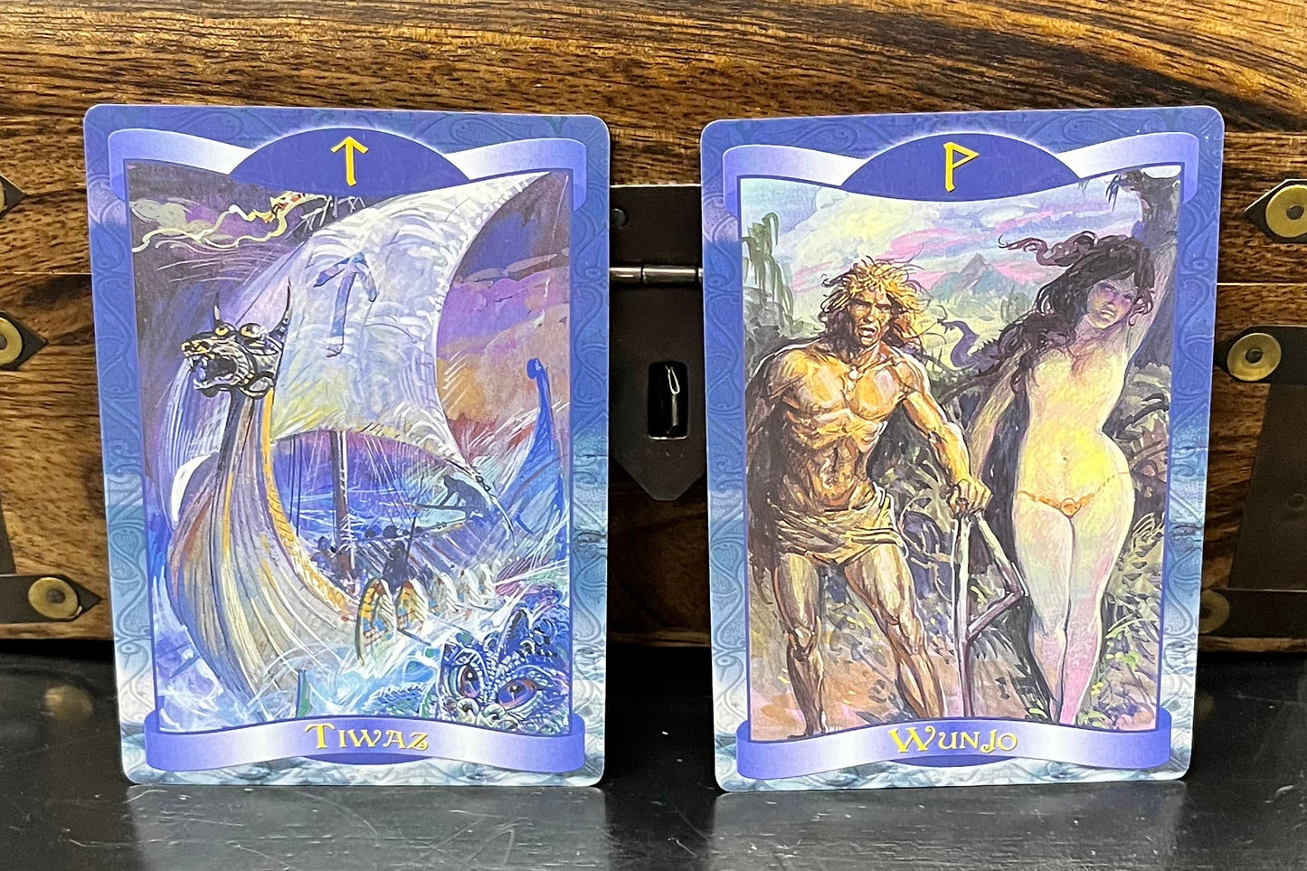 Two illustrated oracle cards with blue borders are displayed in front of a wooden chest on a black table.
