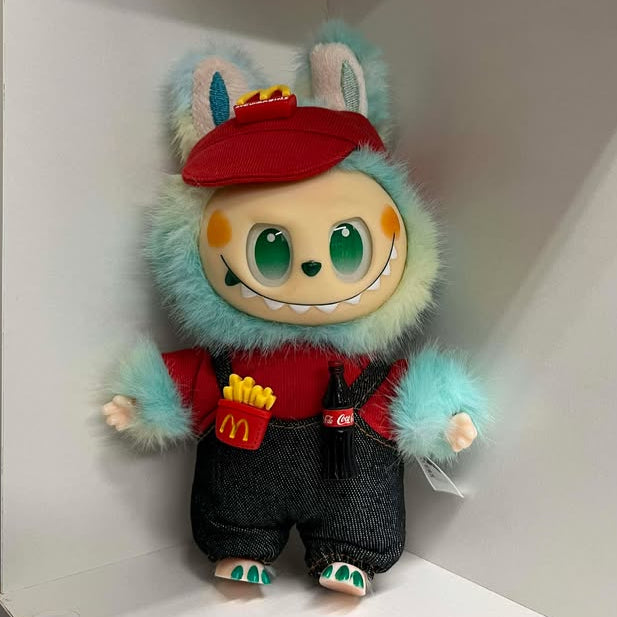 Plush toy with a fuzzy blue design on a white shelf. It is wearing a red visor with the Mcdonalds logo. It also has on black overalls over a red shirt. On the overalls are a plastic container of French fries and a bottle of Coca Cola.