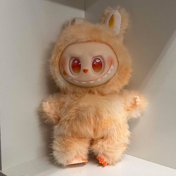 Plush toy with a fuzzy peach color design on a white shelf. It has pink/orange/yellow eyes, an orange nose, a wide smile showing many teeth, a spot of blush on each cheek, and plastic hands and feet. The feet have three toes with painted nails.