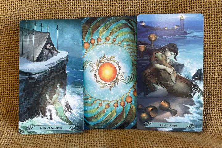 Three tarot cards on a textured brown surface. One depicts a woman in a tent surrounded by swords, on a cliff above a sea from which a ghastly mermaid appears. One is face-down, with a background with a yellow-orange circle in the center, surrounded by a small circle of seaweed, and then by a larger circle of seaweed. The ghird shows a woman on a beach, hugging a seal as three cups lay on the beach beside her and two sit upright behind her.