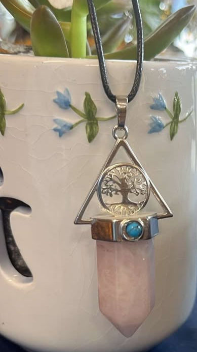 Necklace with a silver colored tree of life pendant surrounded by triangle with blue howlite circular gem inset in the metal and a larger pink crystal. The necklace is held against a white decorative pot.