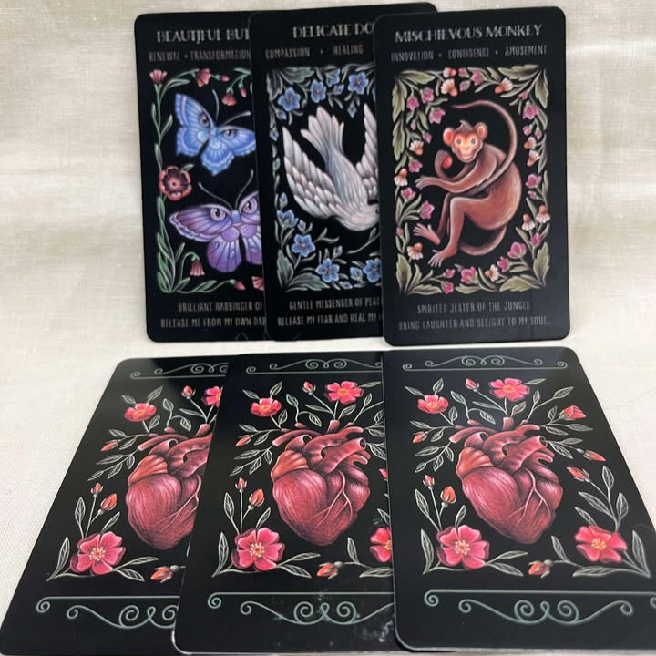 Set of tarot cards with artistic depictions of animals surrounded by flowers on the front and a design of a heart surrounded by flowers on the back on a black background