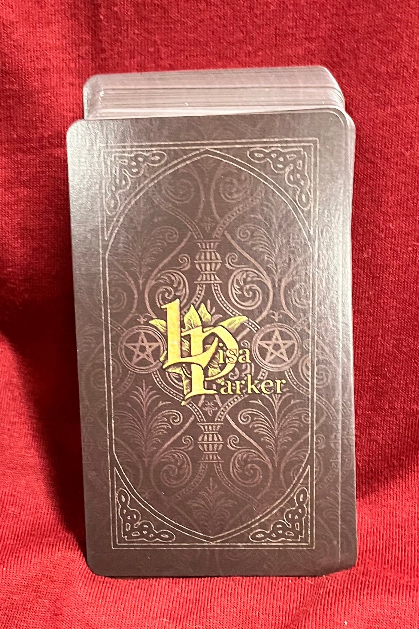 A deck of tarot cards with a dark brown ornate back and a gold 'LP' monogram, resting on a red fabric background.