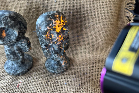 Gray and black patterned stone Jack and Sally statues on a tan textured background. A black light is shining on Sally and parts of the stone are glowing a yellow-orange color.