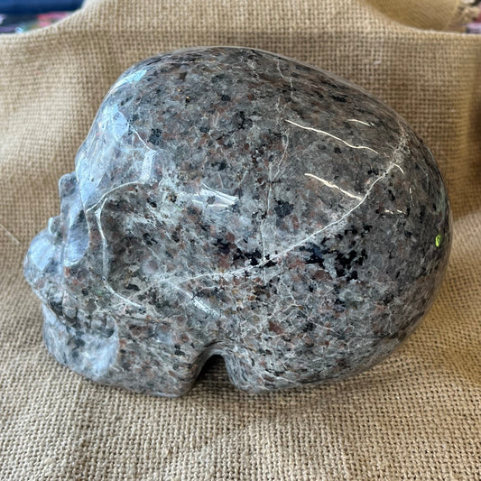 A gray and black patterned stone skull sits on on its side on a tan textured background. Some cracks are visible running through the crystal.