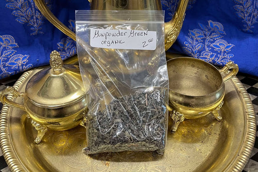 Grayish-green tea leaves in a clear plastic zip bag sit on a golden tea tray surrounded by a tea set.  The bag is labeled "Gunpowder Green tea, organic, $2.50 per oz.