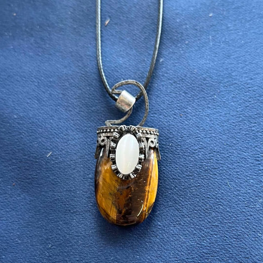 Necklace with a tiger's eye pendant, with a moon outline made from dull metal above it and a white cat's eye stone inset into metal above the tiger's eye on a blue background