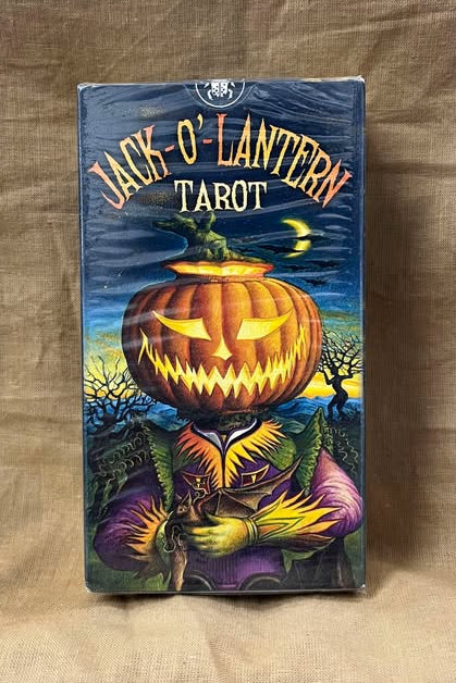 Jack O' Lantern Tarot card deck on a textured surface features a grinning jack o lantern head on a human-esque body, in front of a field with a few trees.