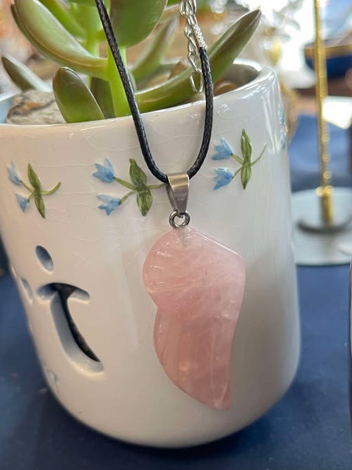Pink rose quartz angel wing crystal  pendant on a black cord in front of a white decorative pot on a blue tablecloth. 