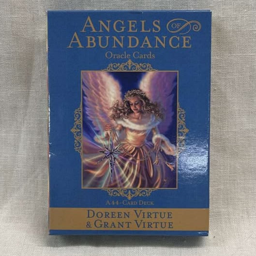 Card deck packaging for 'Angels of Abundance' oracle cards has a blue background with a blonde angel with curly hair, a pink dress, and wings holding a bright star. 
