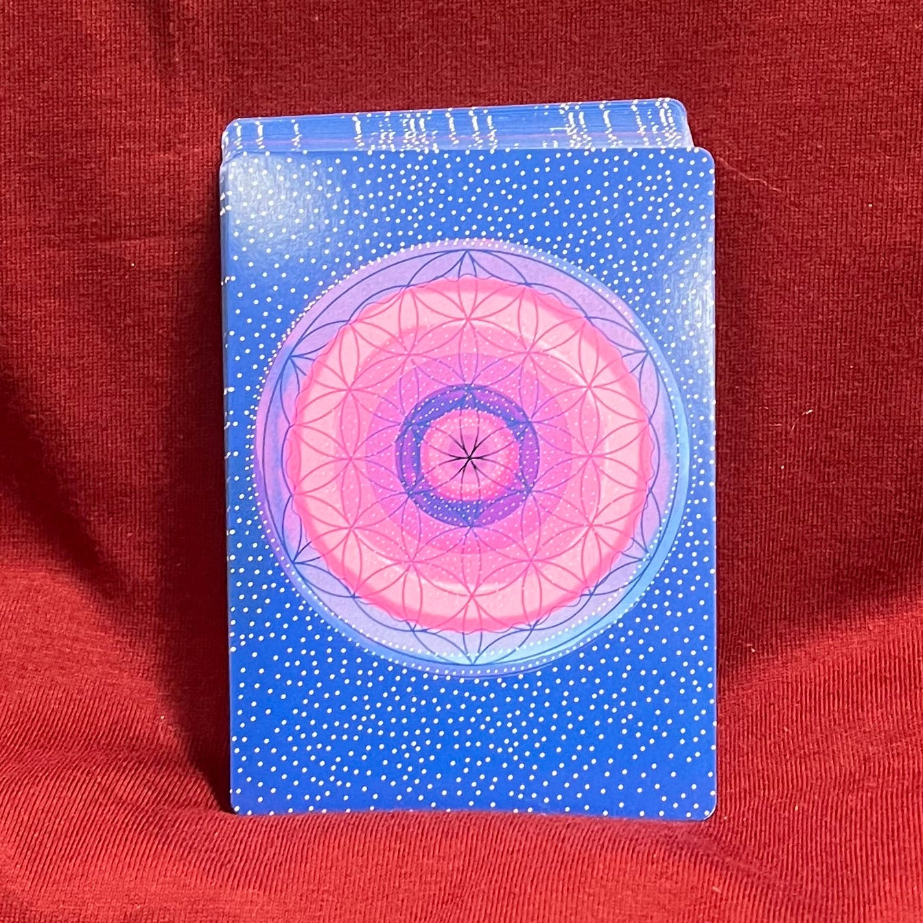 A blue card back with a pink and purple concentric circle design stands upright on a red fabric surface.