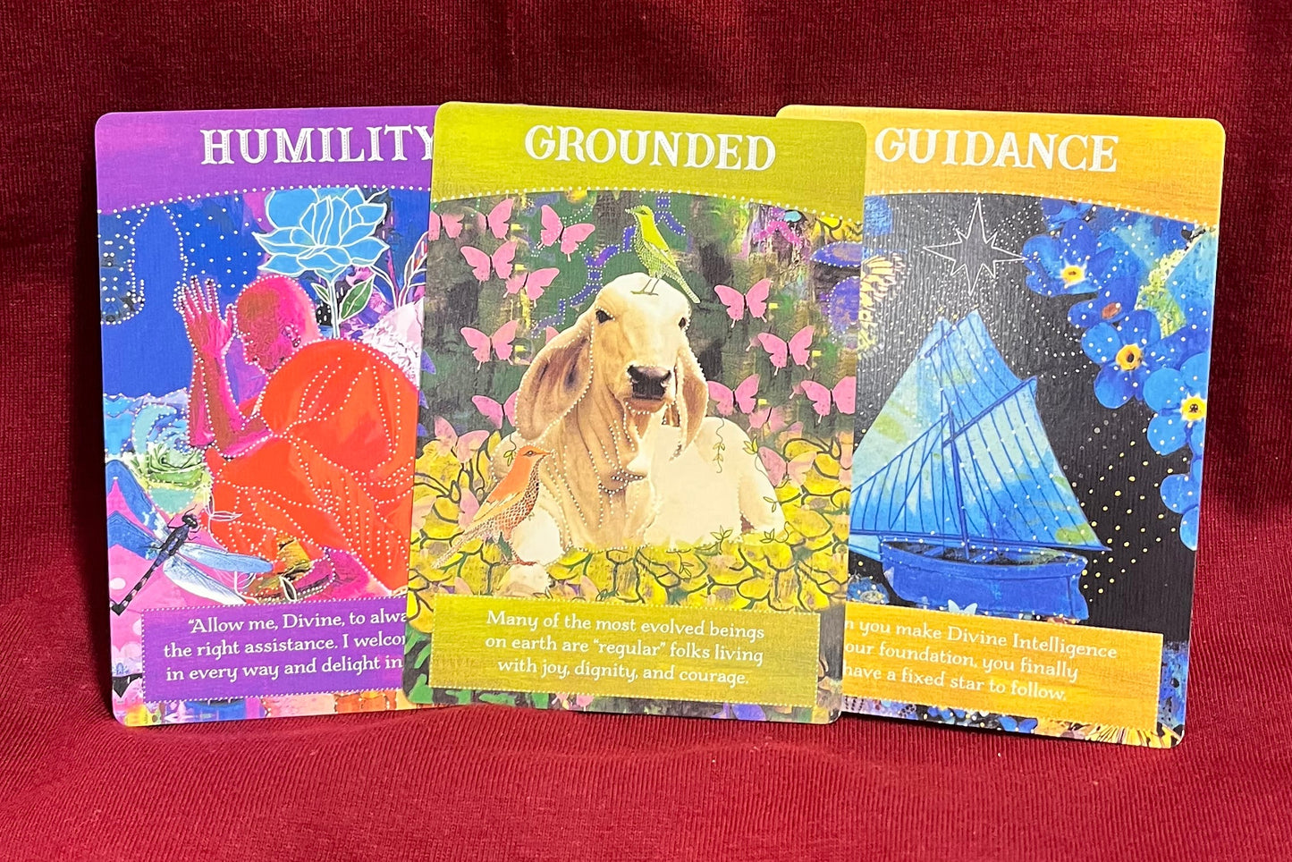 Three colorful oracle cards titled 'Humility,' 'Grounded,' and 'Guidance' displayed on a red fabric background.