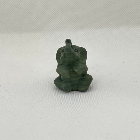 Small green elephant figurine on a white surface. The elephant is sitting down with his trunk in the air.