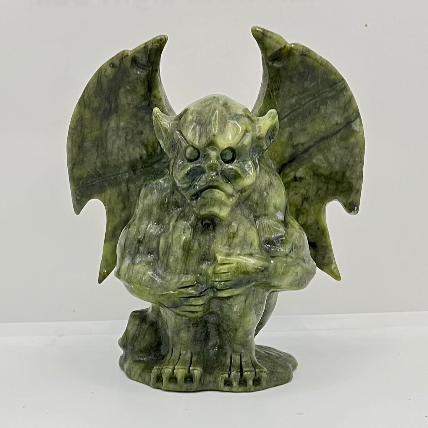 Green jade gargoyle on a white background. The green color is varigated and the gargoyle is clutching his legs to his chest with his wings out.