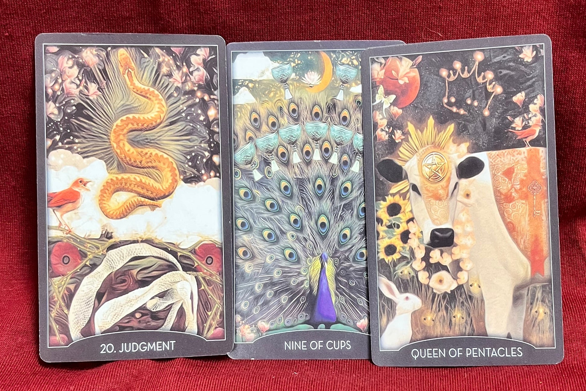 Three illustrated tarot cards are laid side by side on a red fabric background. The visible card titles at the bottom read 20. Judgement, Nine of Cups, Queen of Pentacles.  