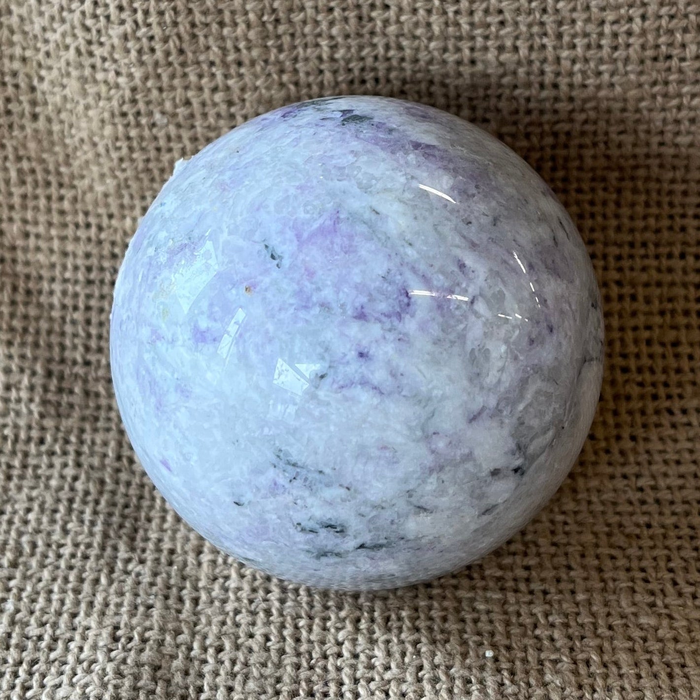 Polished white and light purple crystal sphere with darker purple marbling on a textured beige background