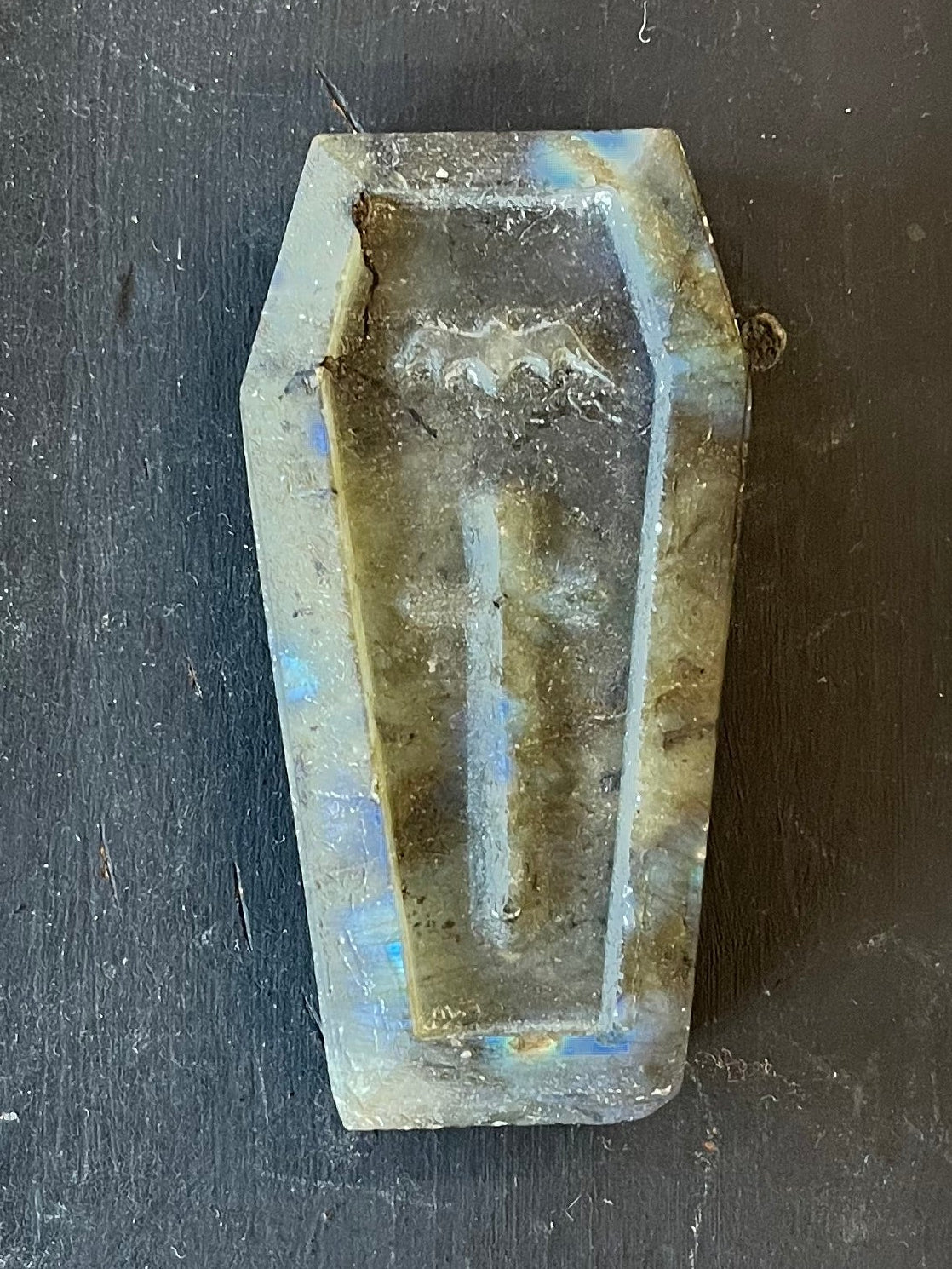 A small coffin-shaped stone carving of  blue-green somewhat irridescent stone placed on a dark wooden workbench. The coffin features a bat and a cross.