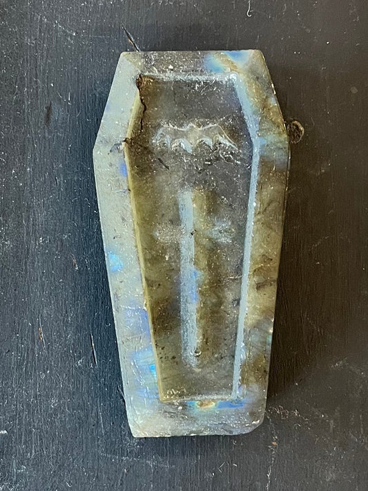 A small coffin-shaped stone carving of  blue-green somewhat irridescent stone placed on a dark wooden workbench. The coffin features a bat and a cross.
