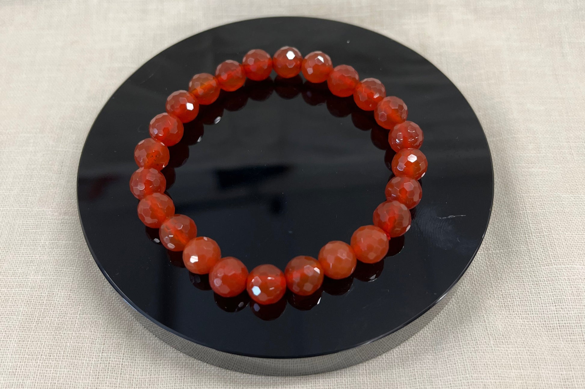 Redish orange faceted carnelian bead bracelet on a black circular stand against a beige fabric background