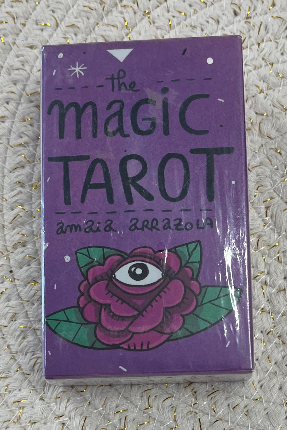 Purple tarot card box with handwritten words "the magic tarot" and "Amaia Arrazola" above a rose design with an eye in the middle.