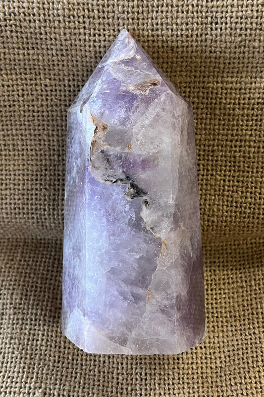 Amethyst crystal on a burlap surface.  The crystal has some brown areas at the top, and some cracks inside with black inclusion on the side. 