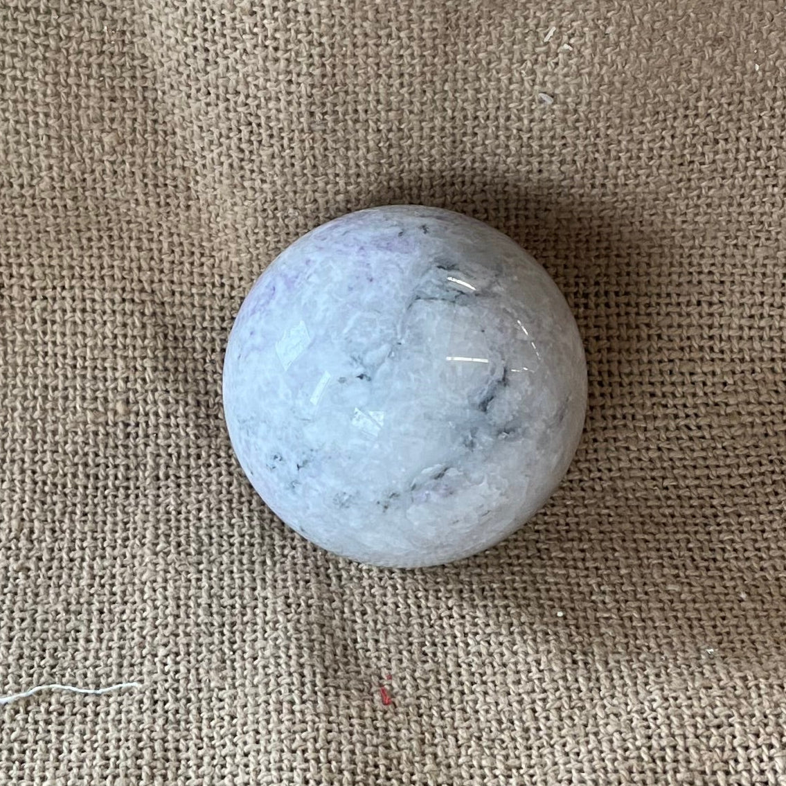 Polished white and light purple crystal sphere with darker purple marbling on a textured beige background