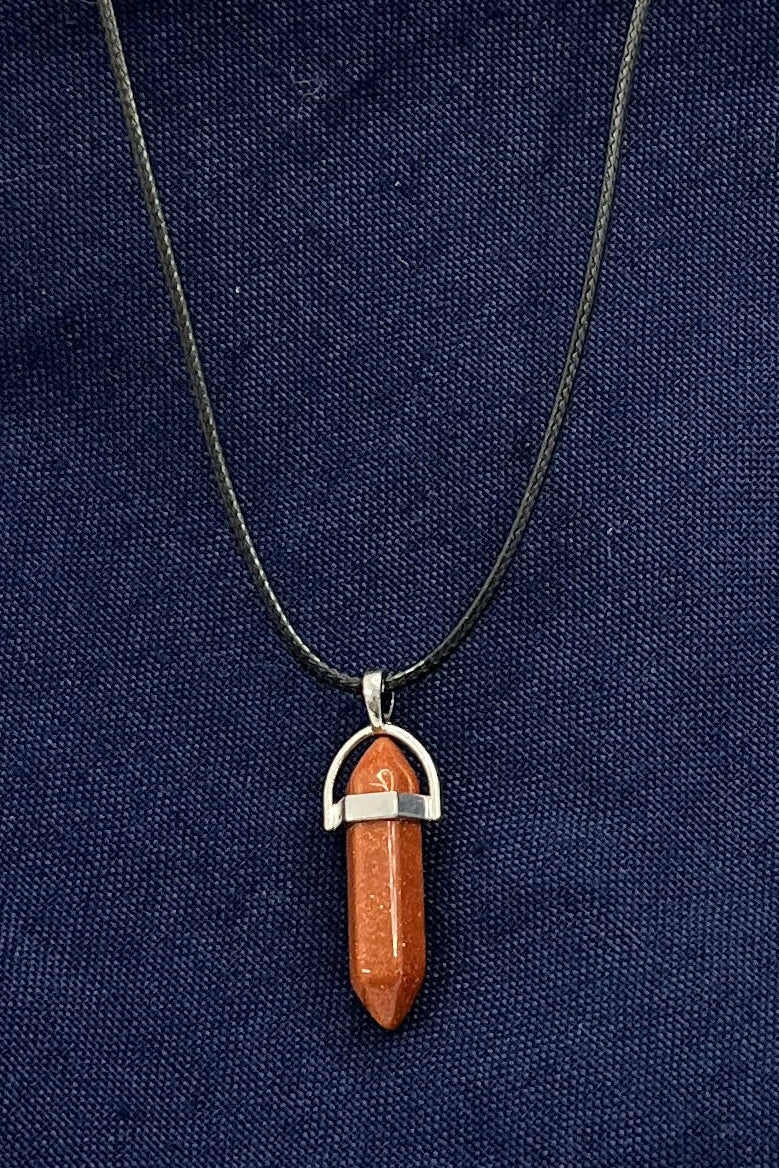 An orange-red sparkling double-pointed gem in a metal finding on a black corded necklace sits on blue fabric