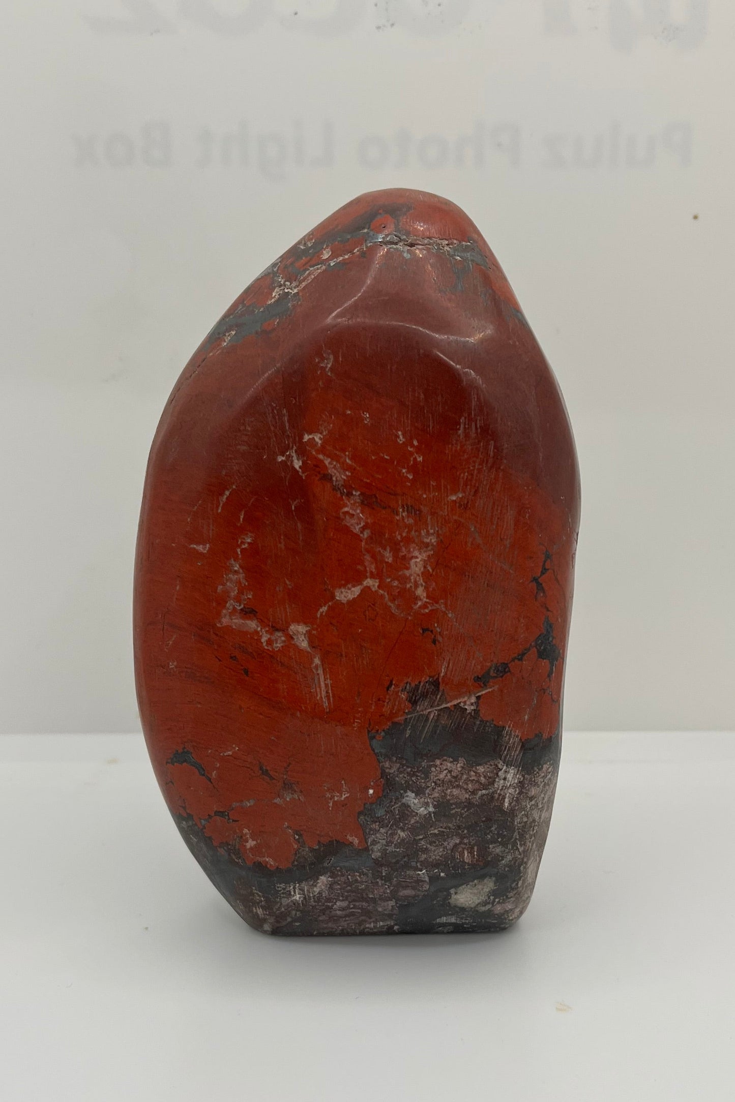 A polished stone of red jasper, red with a bit of white marbling in the top, and areas of darker gray scattered.  The stone is rounded on one side, flat on the other, and has a flat area on the bottom.