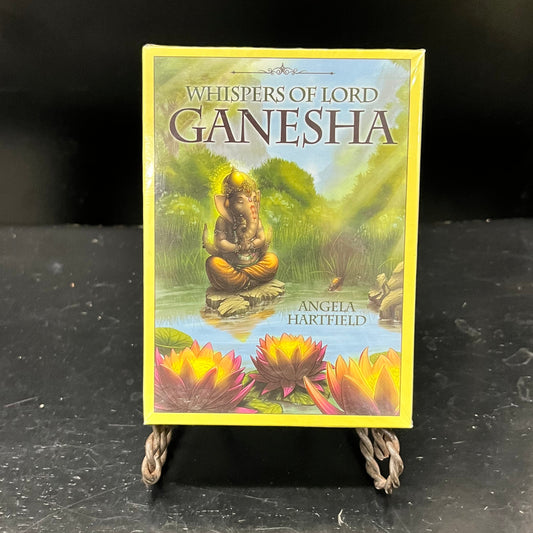 A card deck labeled "Whispers of Lord Ganesha" shows an elephant, in clothing and jewelry, meditating on a rock in the middle of a pond with flowering woods behind it.  Glowing lotus flowers float on the surface of the pond.