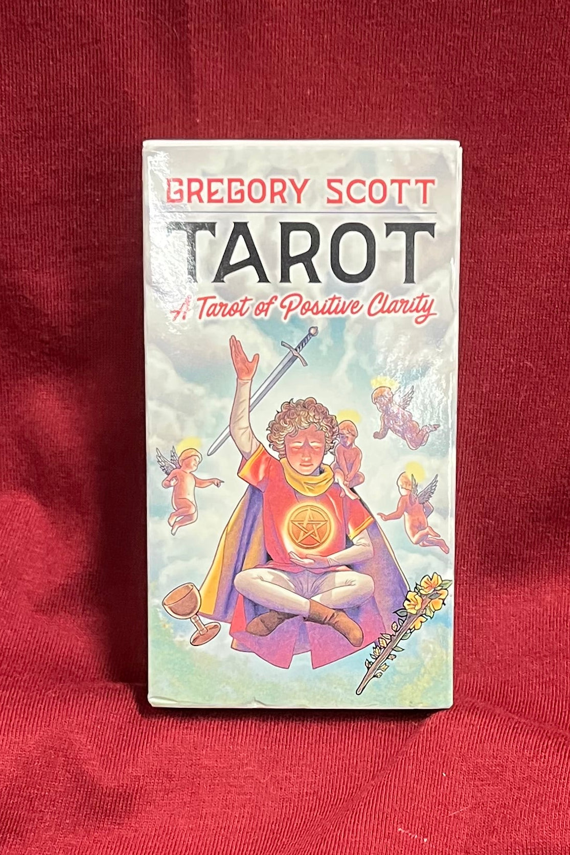 A boxed tarot card deck titled 'TAROT - A Tarot of Positive Clarity' by Gregory Scott stands upright on a red fabric surface.