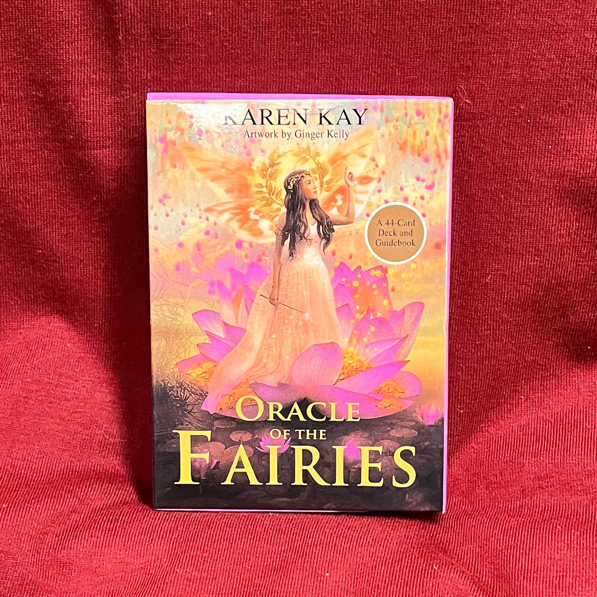 A boxed card deck titled "Oracle of the Fairies" by Karen Kay stands upright on a red fabric surface. The box features fantasy artwork with pink, orange, and purple tones, with a beautiful fairy. 